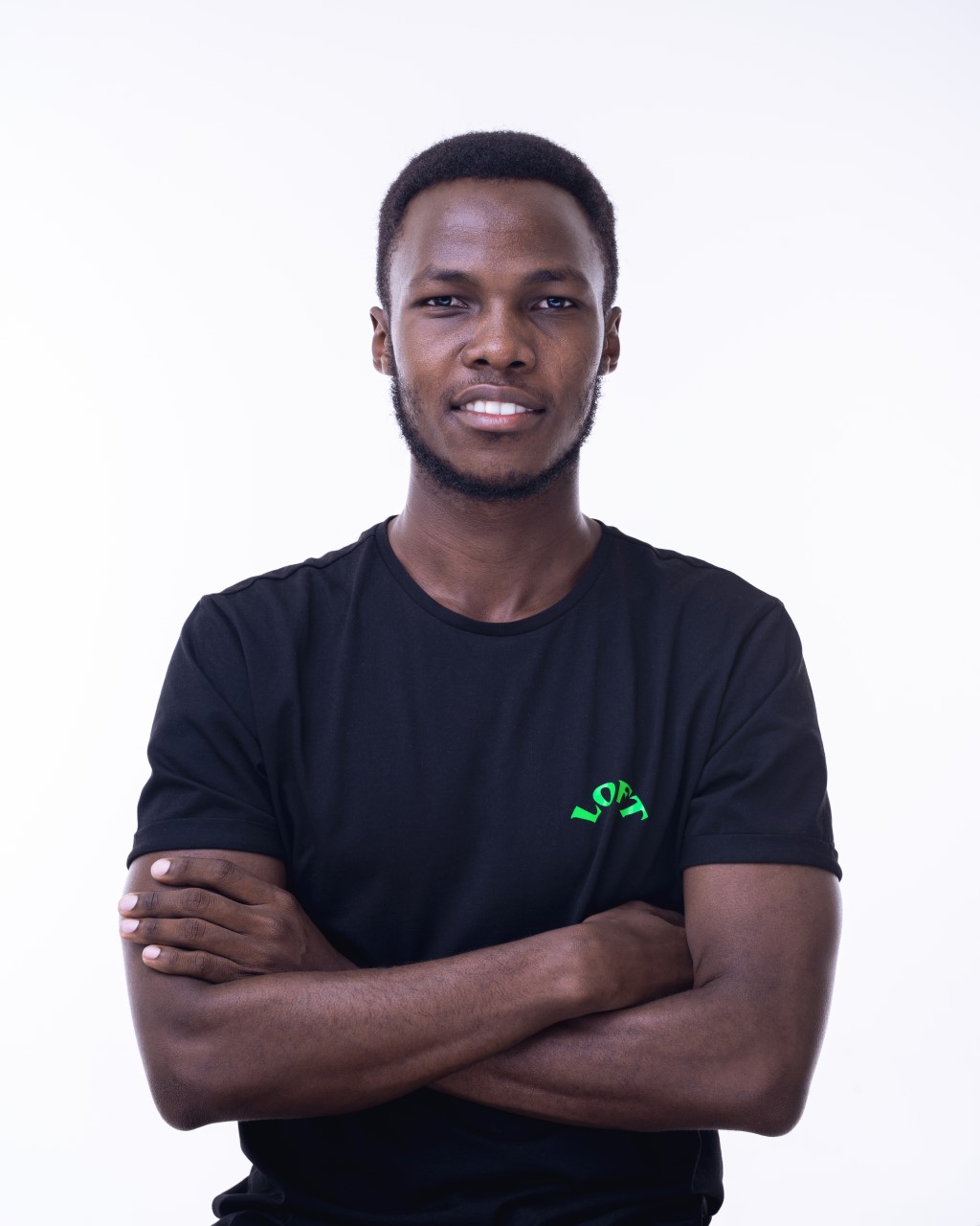 Akbr Kanyesigye, arms folded, smiling, in a black T-shirt against a white wall